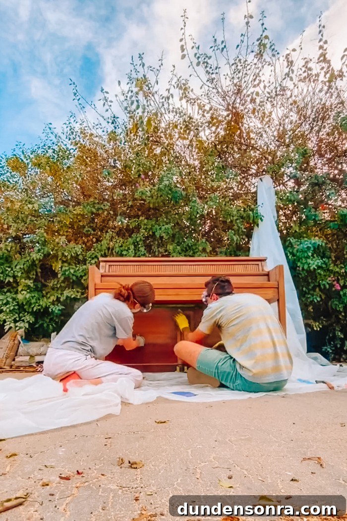 Man and woman sanding a wood piano outside in front of greenery