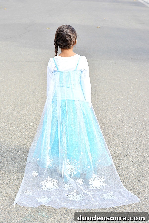 Elsa Christmas Costume Girl standing with her back to the camera, dressed with a blue dress and long white veil with snowflakes on it.