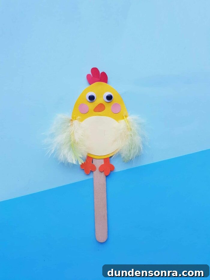 Chick made out of construction paper and glued to a wooden stick on a blue background. 