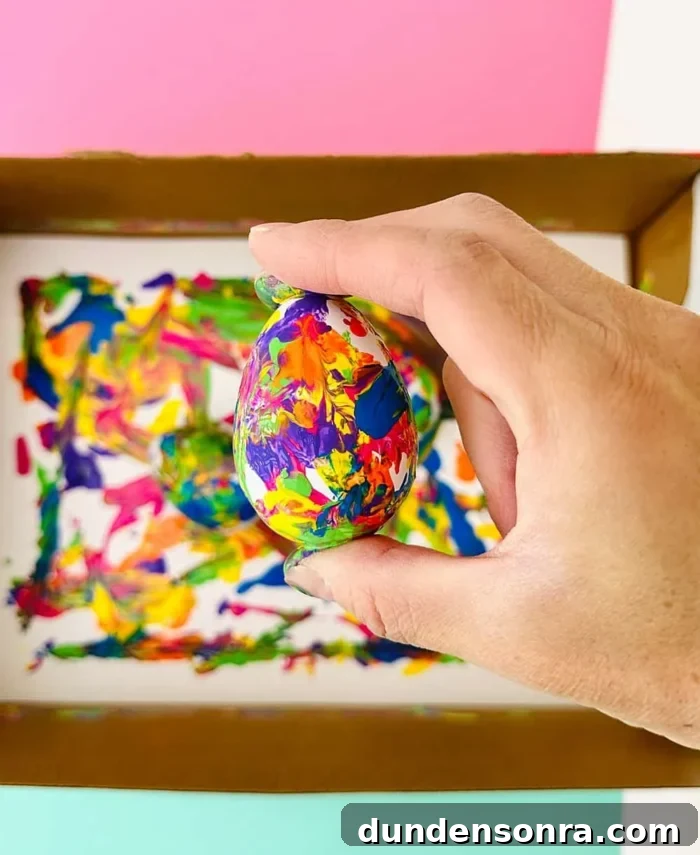 Hand holding an egg covered in paint with a cardboard box full of paint in the background. 